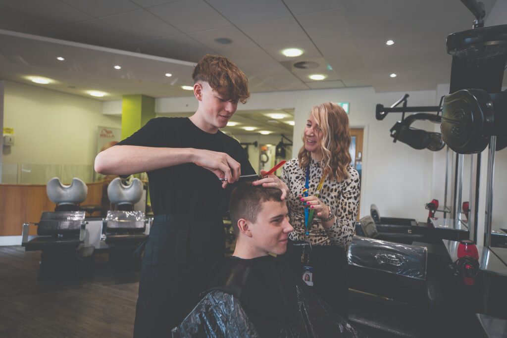 A barbering student practicing cutting hair on somebody with a tutor stood to their left