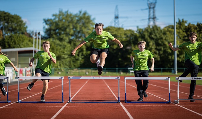 Five sports students in green running on the Deeside athletics track with the middle student in mid air over a hurdle