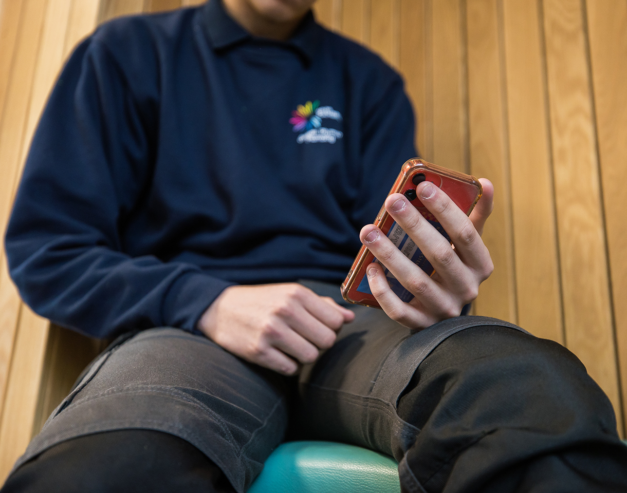 A student sitting down holding their phone wearing a Coleg Cambria jumper