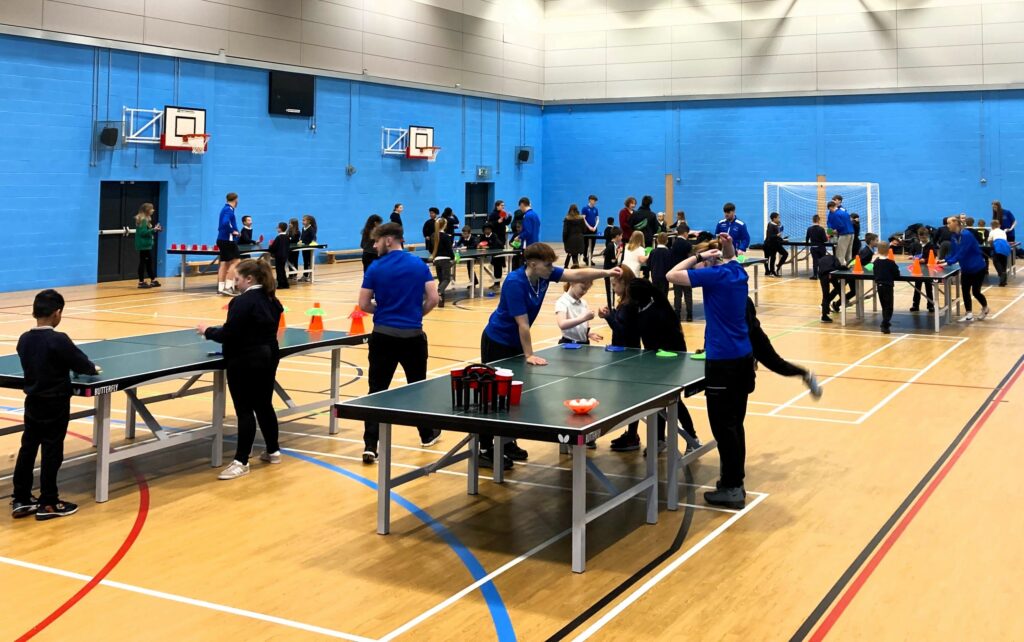 Students and young kids in a sports hall playing table tennis, the image is for the news post 'Coleg Cambria rallied behind a popular tennis academy which went from strength to strength in 2024'