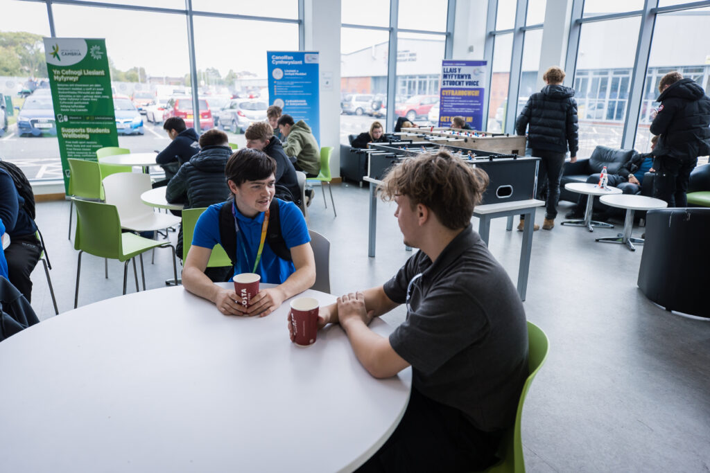 two students sat at a table drinking a hot drink whilst looking at eachother