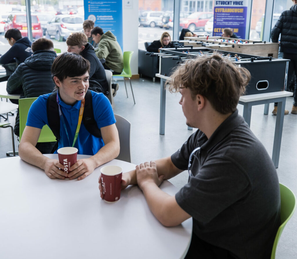 two students sat at a table drinking a hot drink whilst looking at eachother