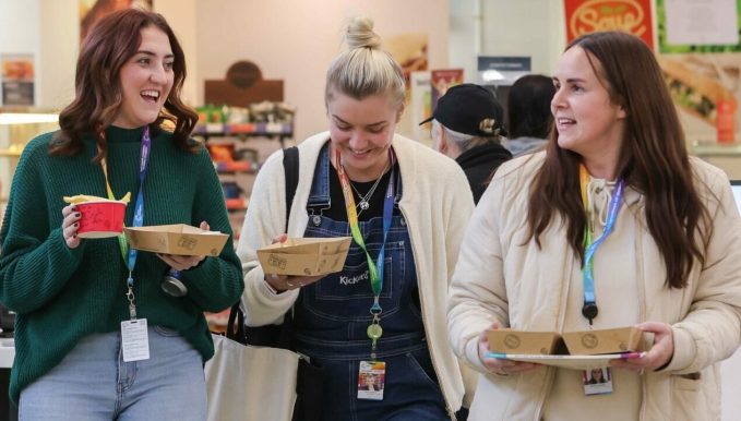 Three students in the canteen walking towards the camera with their breakfast